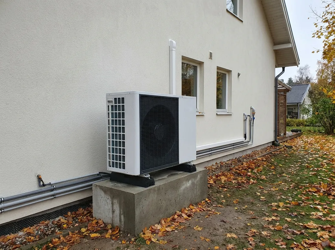 Jednostka zewnętrzna pompy ciepła powietrze-woda przy ścianie domu jednorodzinnego, obok przewody w izolacji prowadzone do wnętrza budynku.